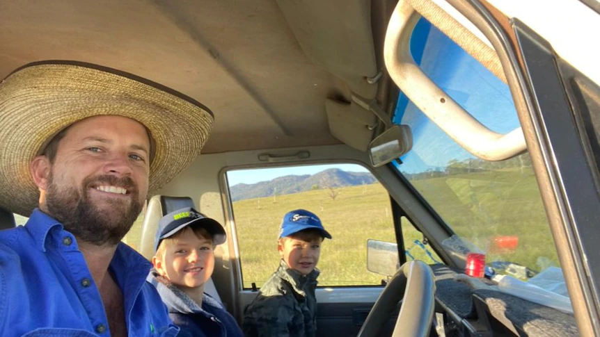A man in a hat and a blue shirt takes a selfie at the wheel of a stopped car, with his two boys beside him.