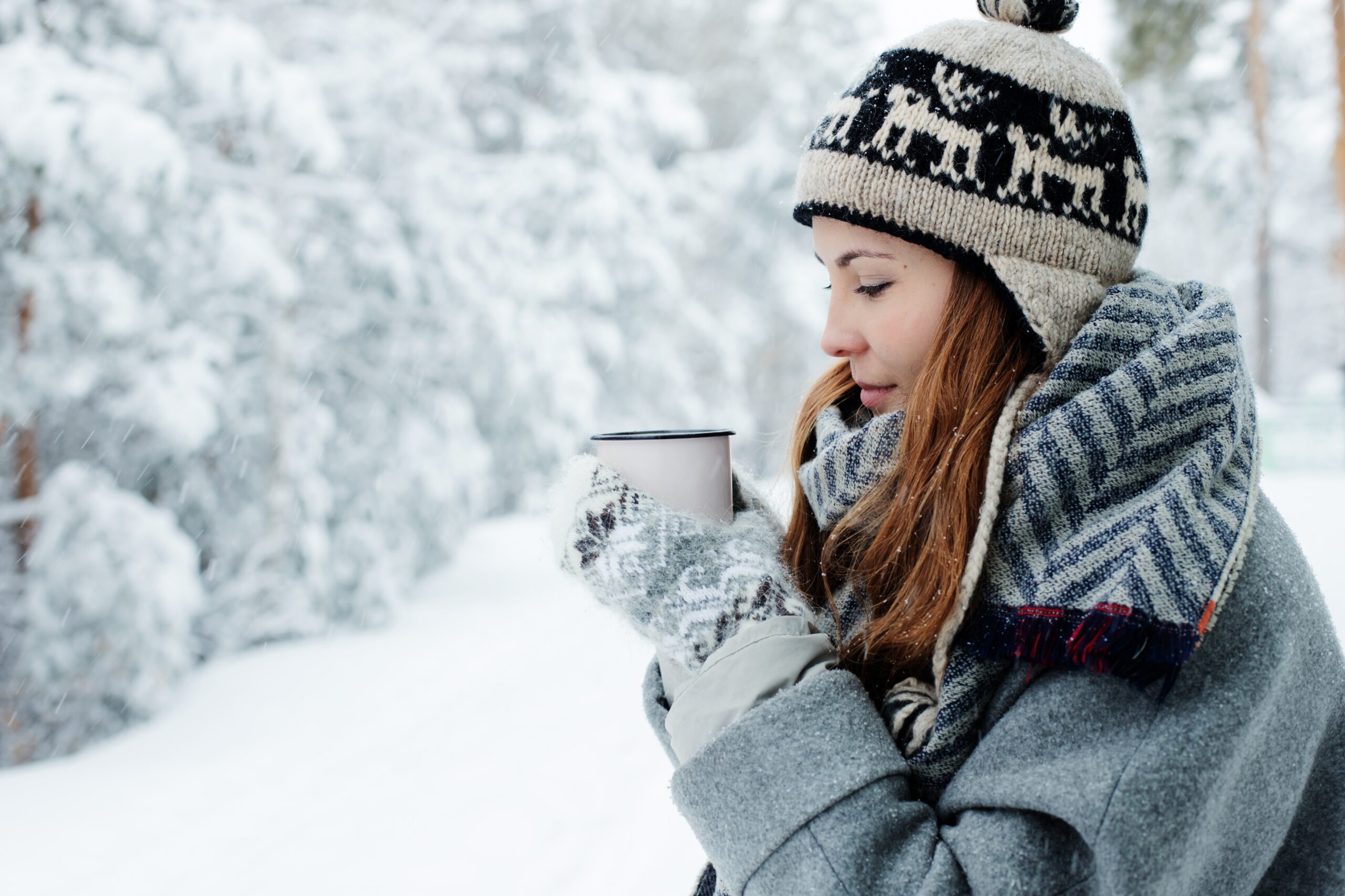 Woman dressed in jacket, mittens, and hat holding a mug outside on a snowy day.